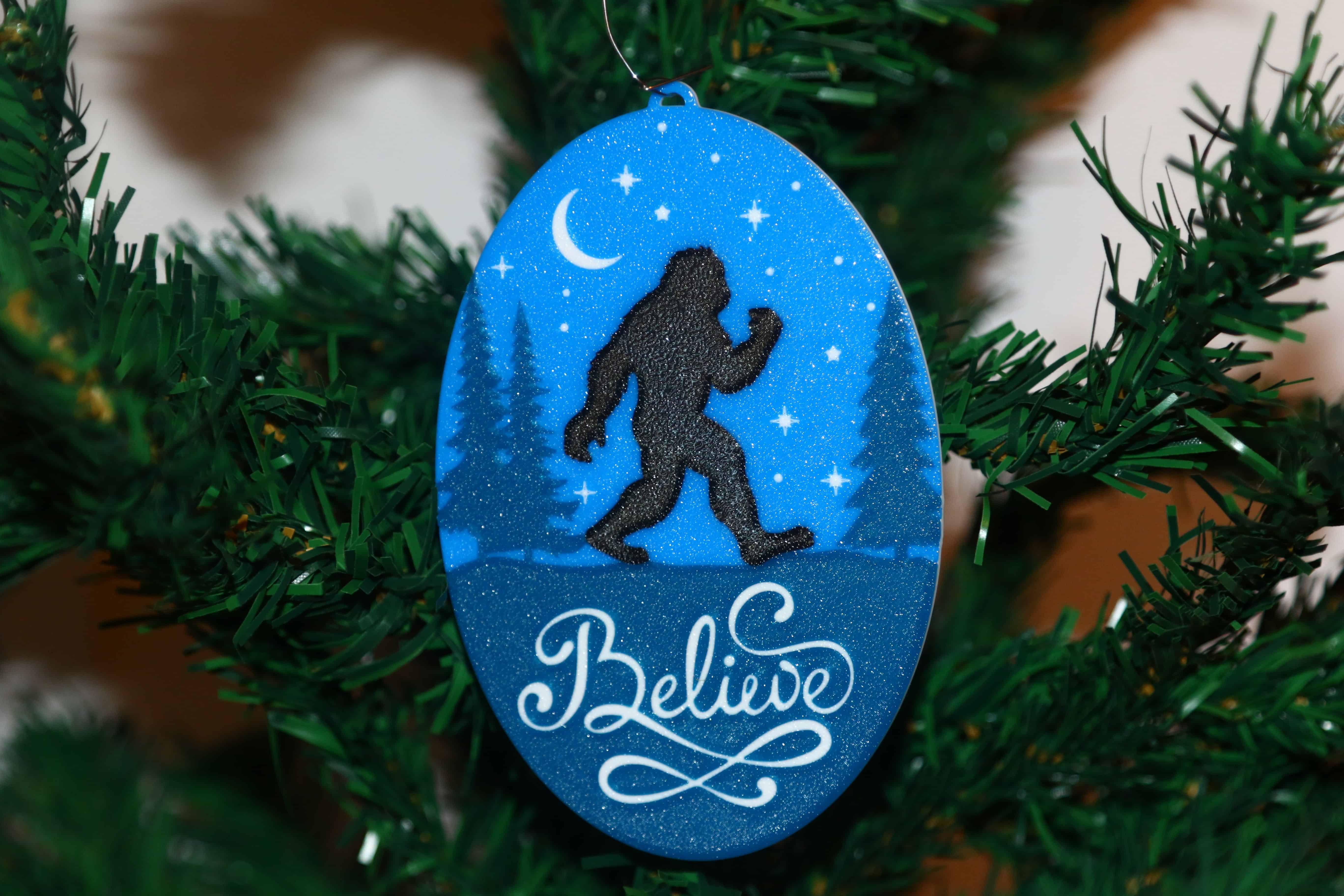 Bigfoot Christmas ornament with forest background, silhouette, and “Believe” text.