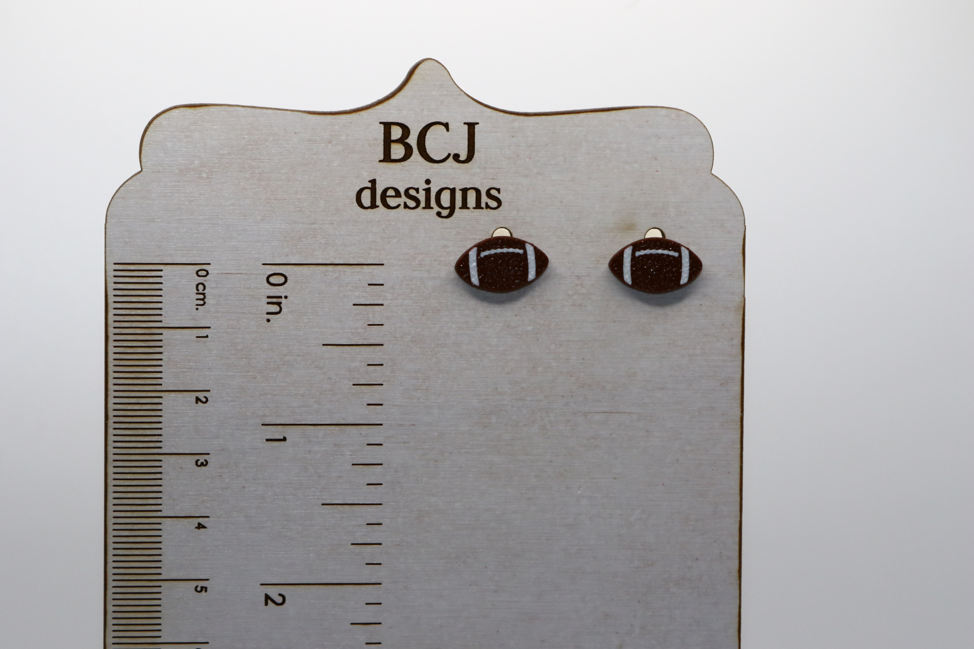 Pair of small stud earrings shaped like a brown football with white stitching.